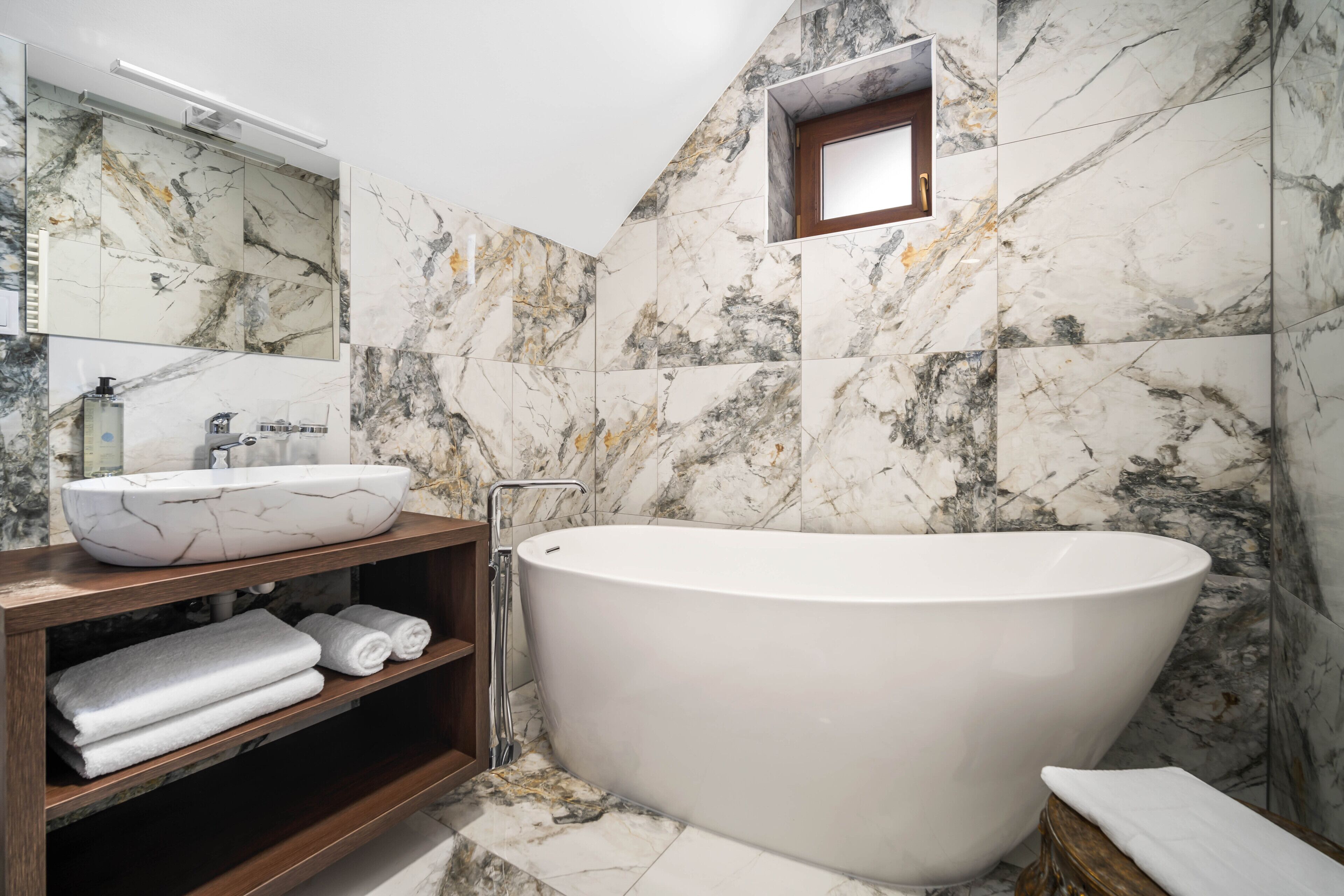 A modern bathroom with a freestanding bathtub, a marble-tiled wall, and a wooden vanity with towels.