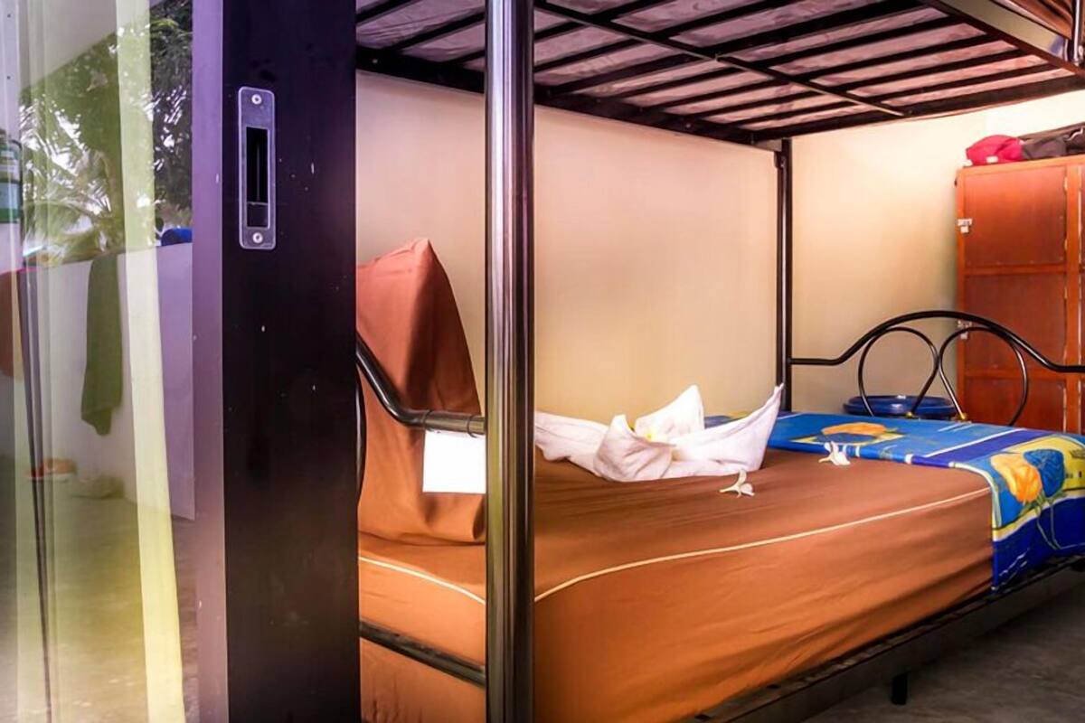 A single bed with a metal frame, a wooden headboard, and a colorful bedspread. A red bag is placed on the bed, and a white towel is draped over the side.