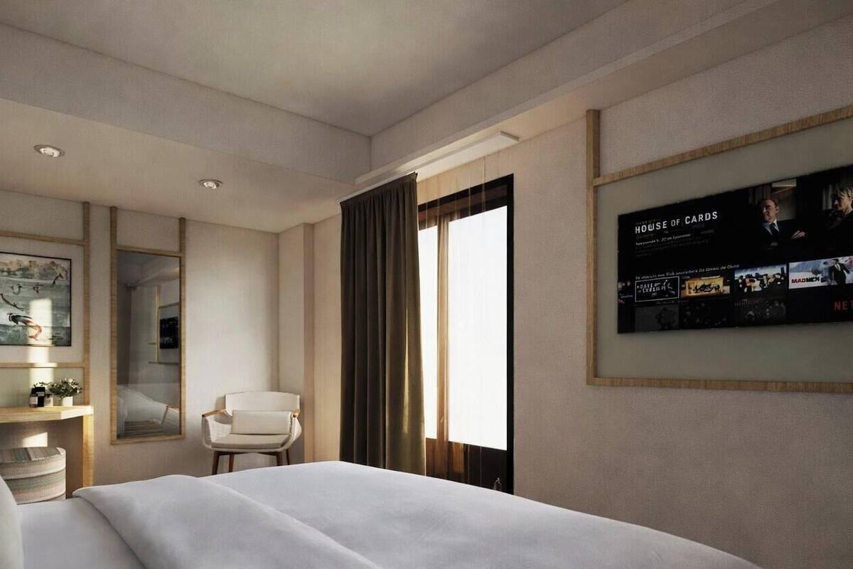 A hotel room with a large bed, a chair, a desk with a lamp, and a wall-mounted TV displaying 'HOUSE OF CARDS'.