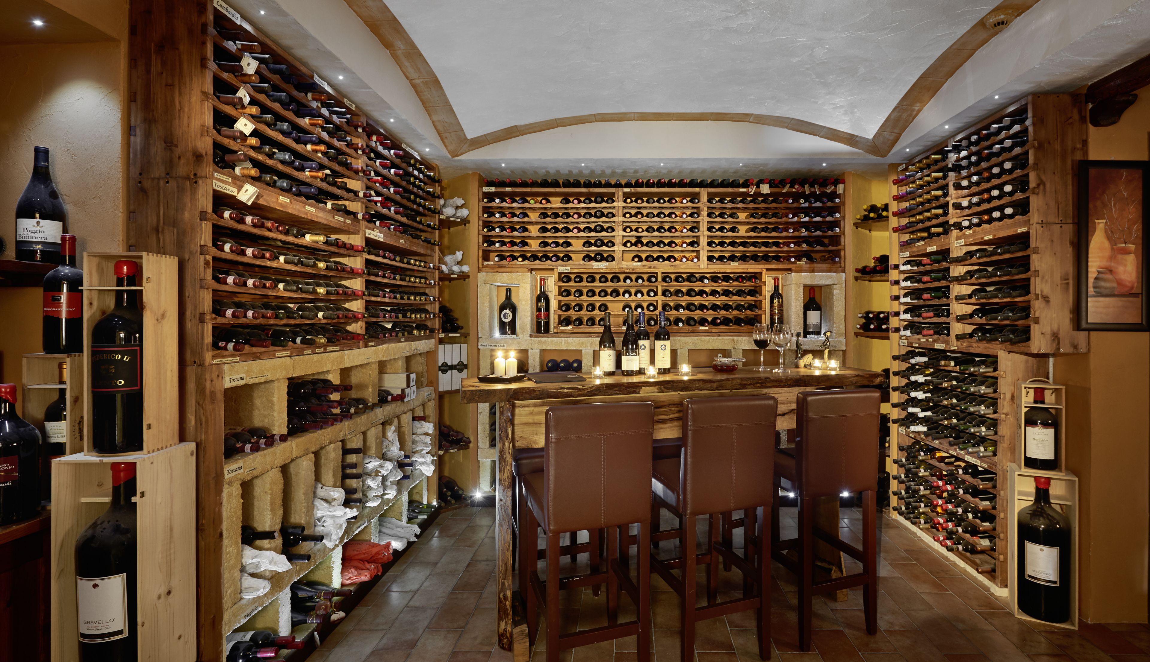 Una bodega con estantes de madera llenos de botellas, una zona de bar con sillas y un suelo de baldosas.