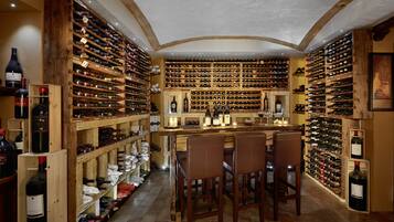 Una bodega con estantes de madera llenos de botellas, una zona de bar con sillas y un suelo de baldosas.