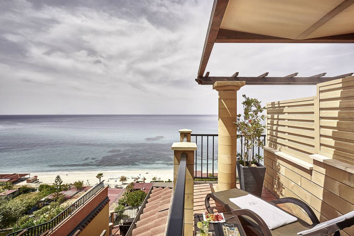 A balcony with a view of the ocean, a table with a drink, and a chair.