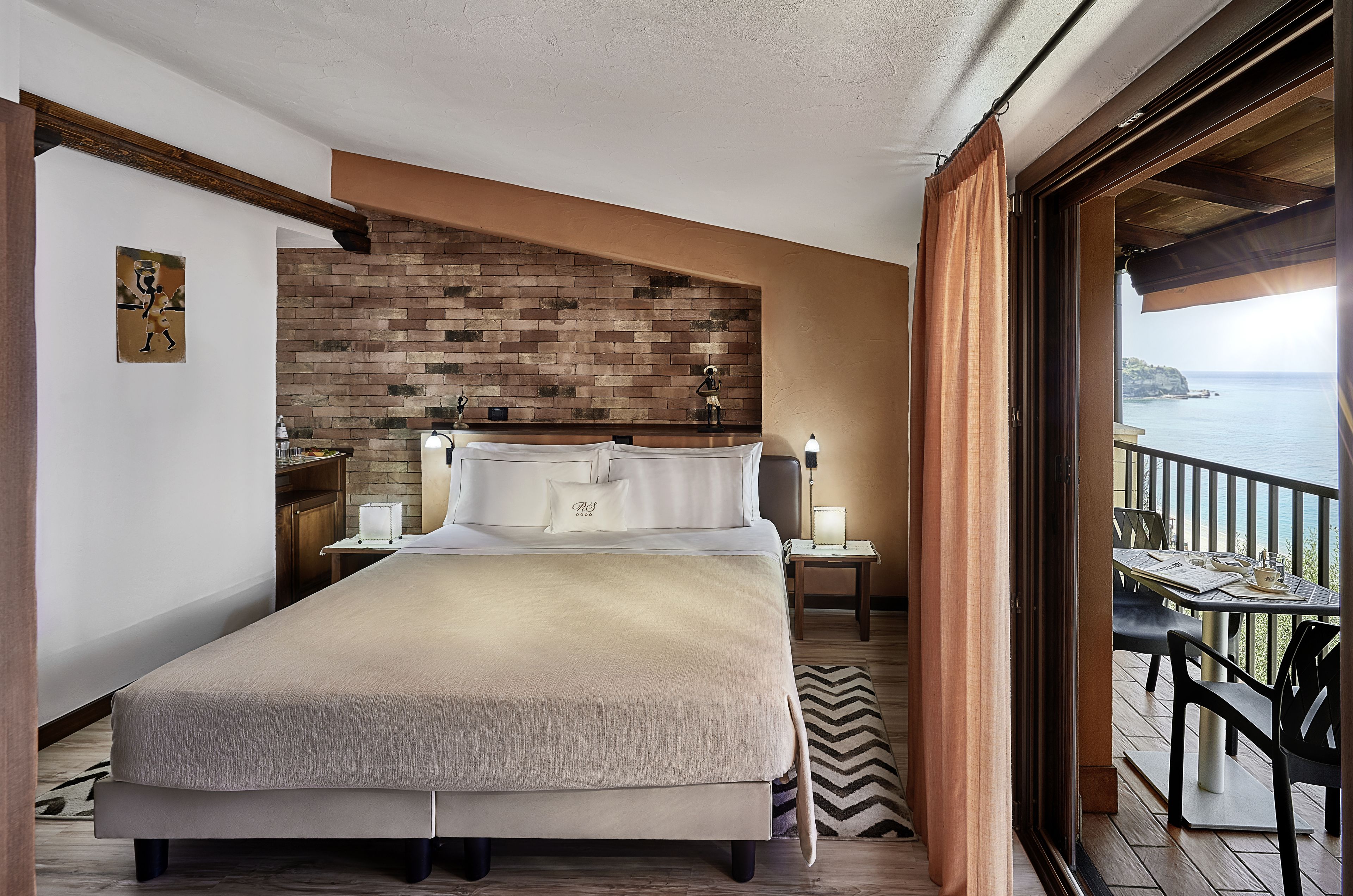 A hotel room with a large bed, a brick accent wall, a view of the sea, and a balcony with outdoor seating.