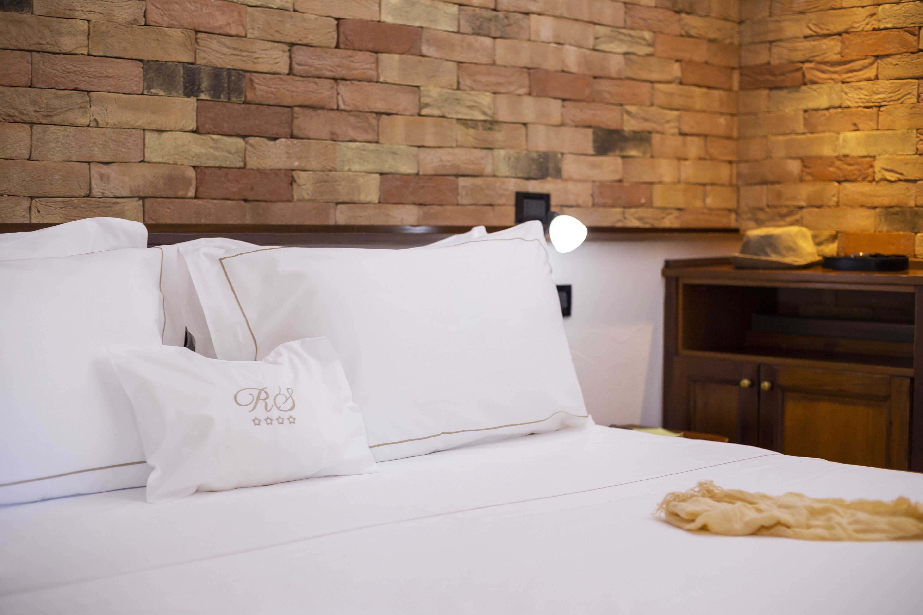 A neatly made bed with white linens and pillows, a brick wall, and a wooden cabinet in the background.
