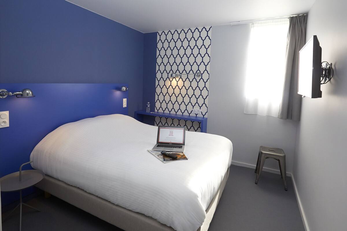 A neatly made bed with a laptop and a magazine on it, a small round table with a lamp, and a window with curtains.