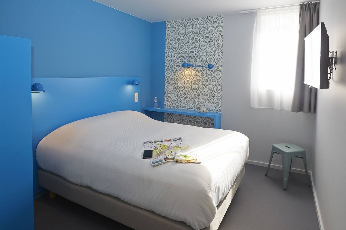 A bedroom with a large bed, a nightstand, a wall-mounted TV, a window with curtains, and a small stool.