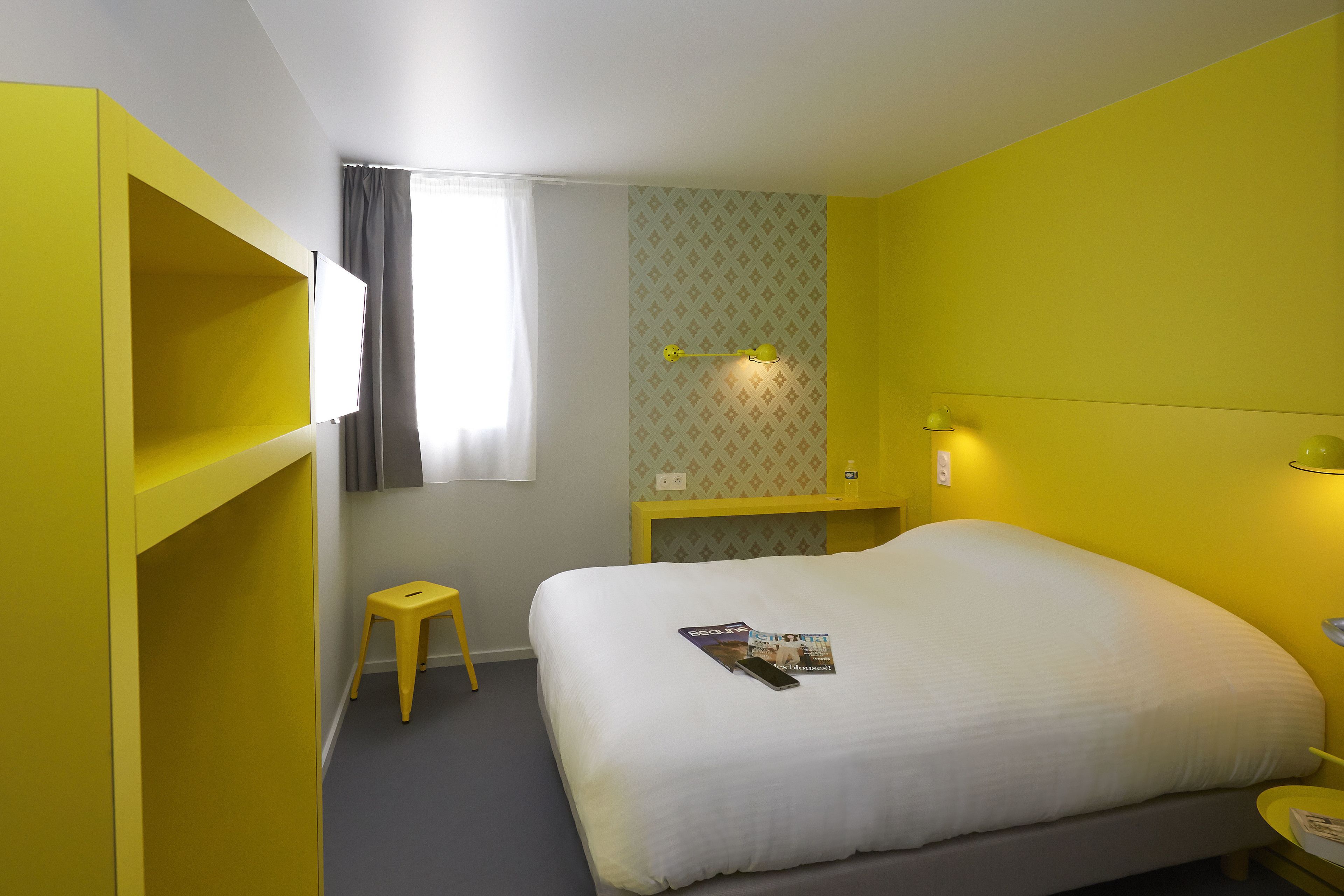 A hotel room with a bed, a small stool, a nightstand, and a window with curtains.