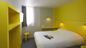 A hotel room with a bed, a small stool, a nightstand, and a window with curtains.
