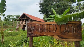 A wooden sign with the text "Luk Tented Camp" in a rustic style, surrounded by lush greenery and a wooden structure with a red roof in the background. - Lak Tented Camp (Lak)