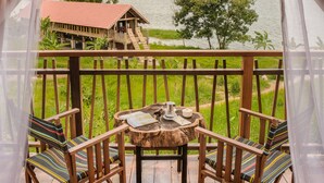 A wooden deck with two striped chairs and a small table, overlooking a lake with a wooden house and mountains in the distance. - Lak Tented Camp (Lak)