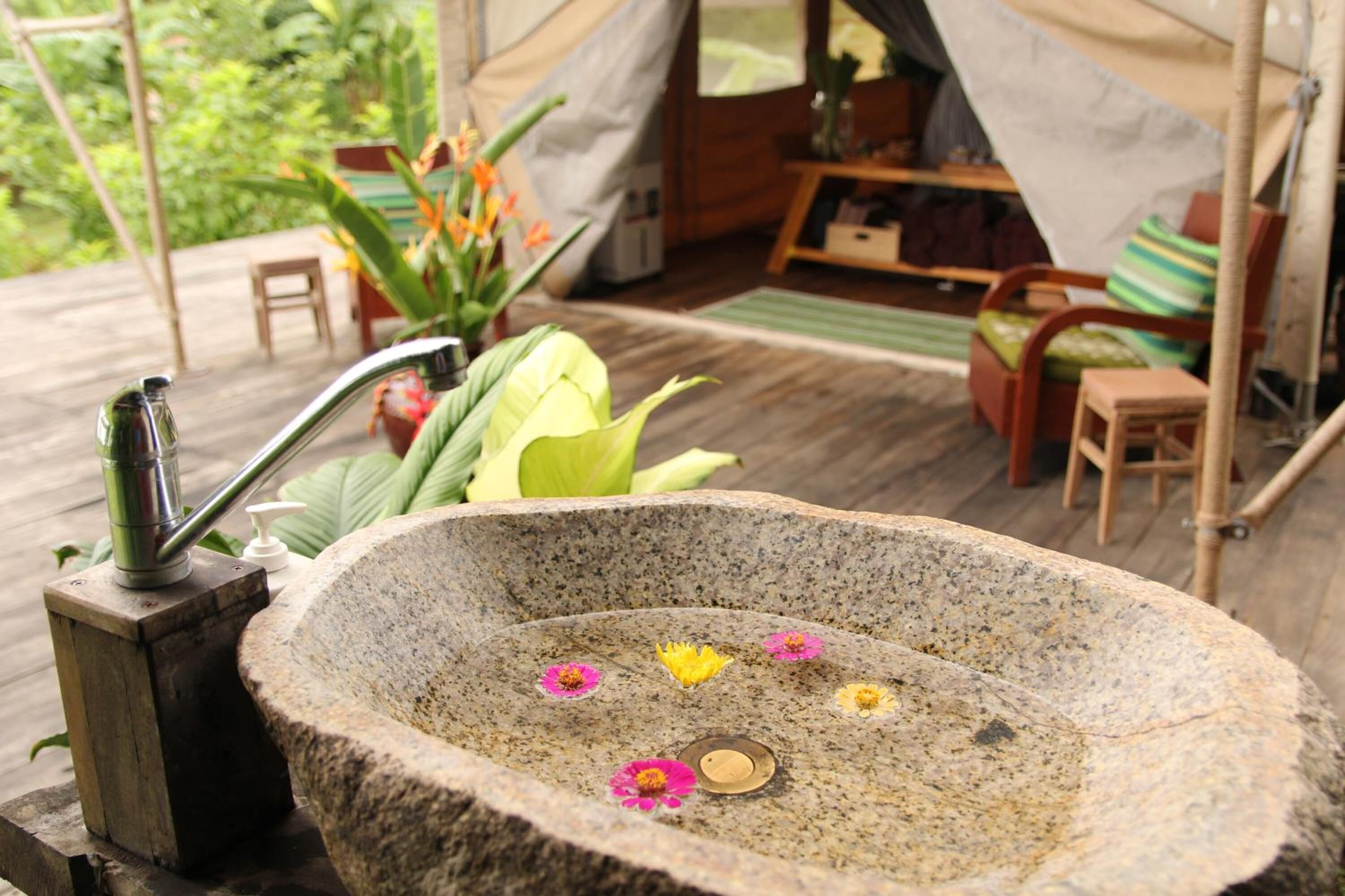A stone basin with flowers and a hand pump, surrounded by wooden furniture and a tent in a garden setting.