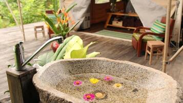A stone basin with flowers and a hand pump, surrounded by wooden furniture and a tent in a garden setting.