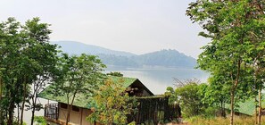 A rustic cabin with a thatched roof, surrounded by lush greenery and overlooking a calm lake with distant mountains. - Lak Tented Camp (Lak)