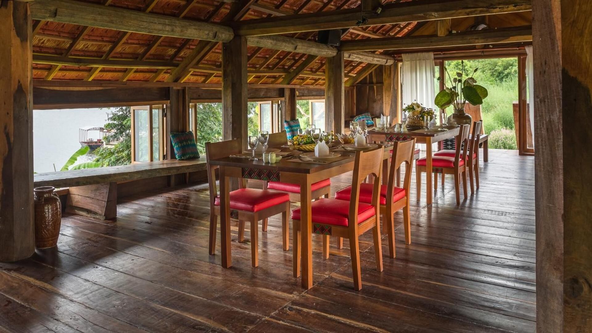 A wooden dining area with red cushioned chairs, a table set for dining, and large windows offering a view of greenery.