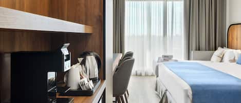A hotel room with a wooden desk, a coffee maker, a chair, a bed with a blue bedspread, and a view of the outdoors through sheer curtains.