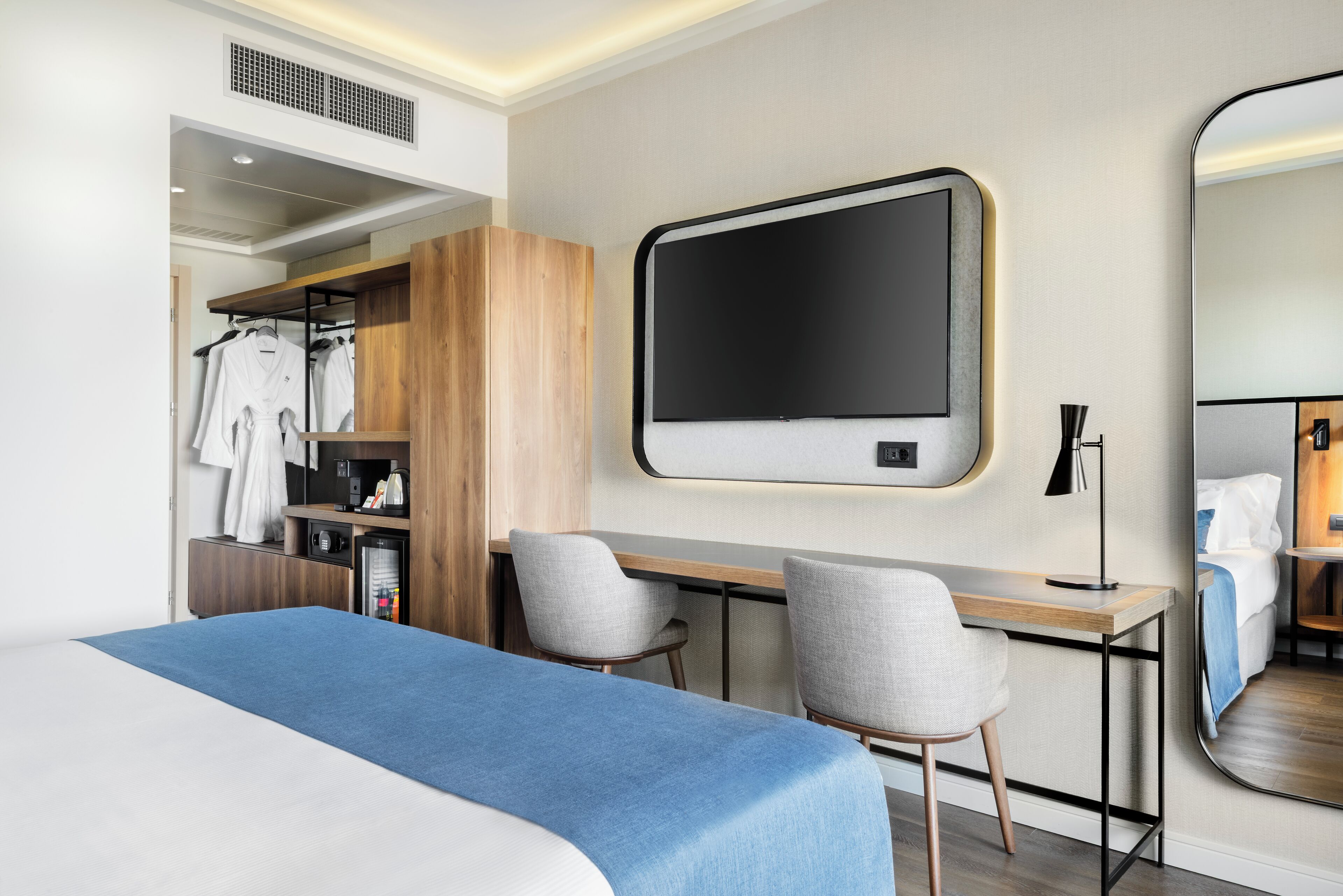 A modern hotel room with a large bed, a desk with two chairs, a wardrobe, a mirror, and a TV.