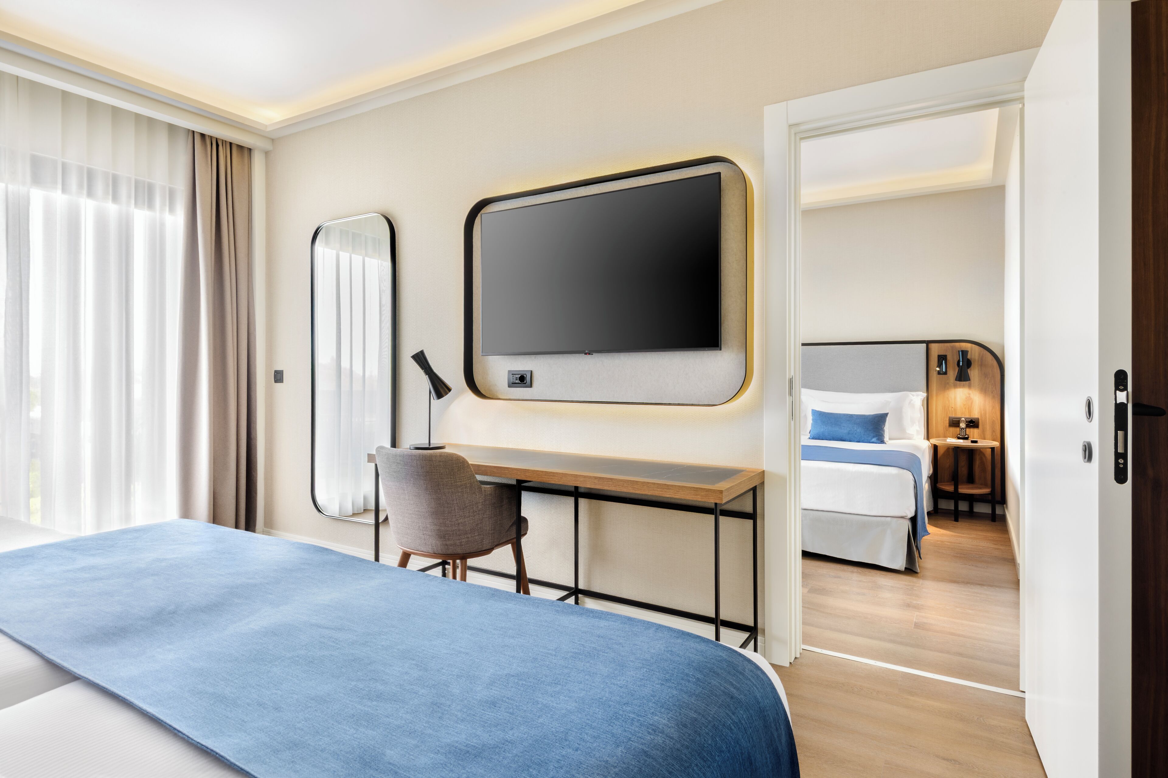 A hotel room with a bed, a desk, a chair, a TV, and a separate bedroom with a bed and a nightstand.