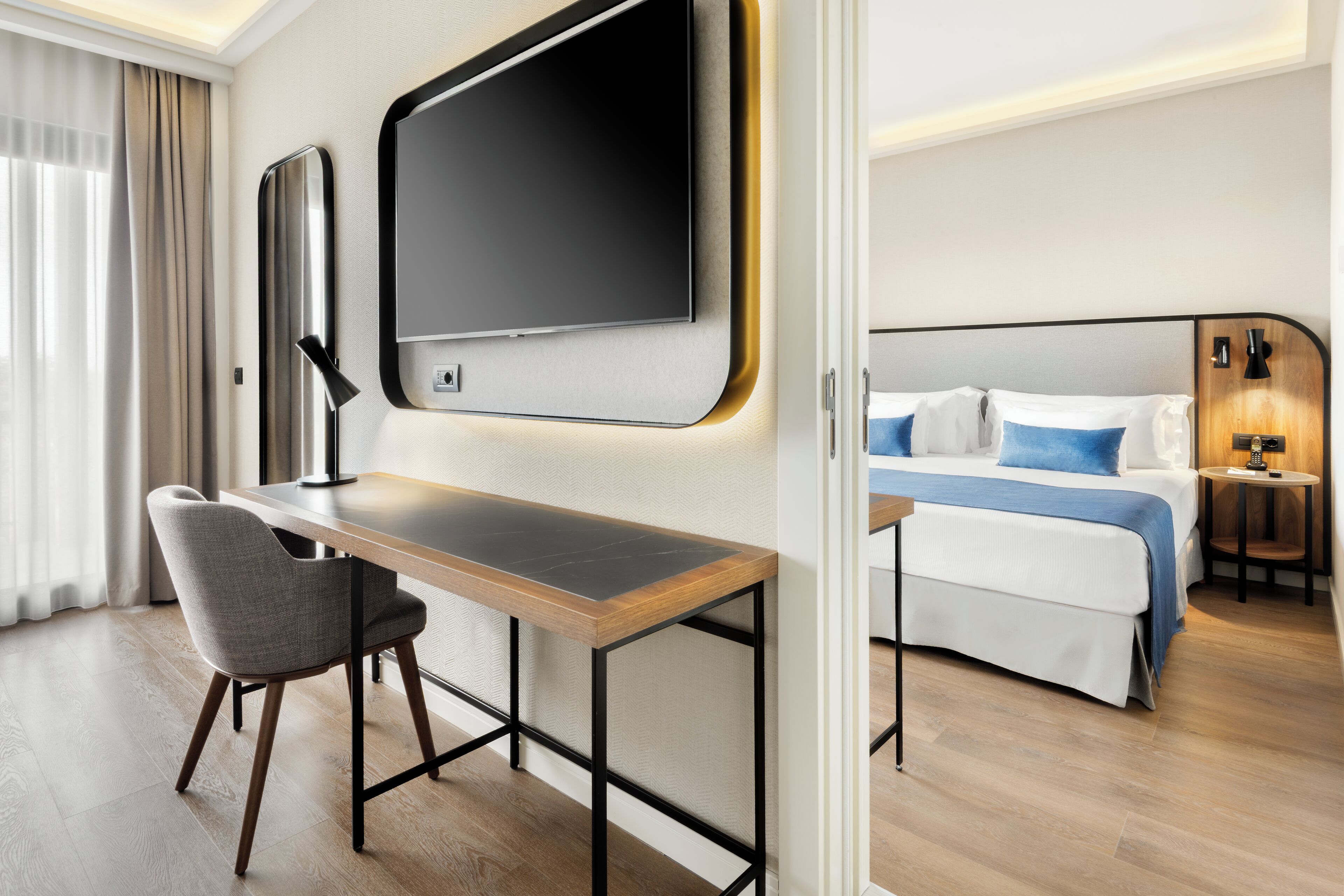 Habitación de hotel moderna con un escritorio de madera, una silla cómoda, una cama grande con ropa de cama azul y un televisor de pantalla plana montado en la pared.