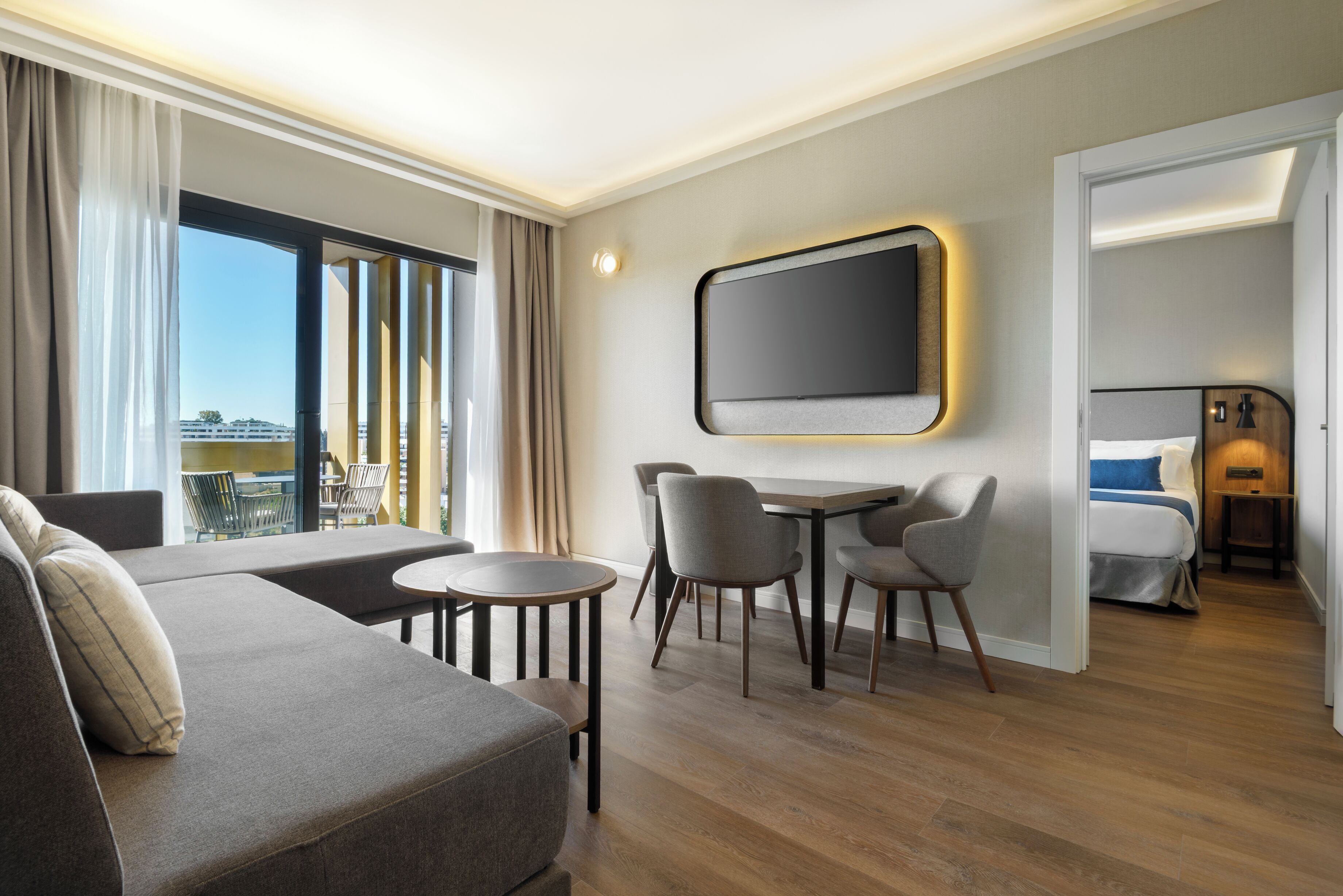 A modern hotel room with a sofa, dining table, chairs, a TV, and a bed.