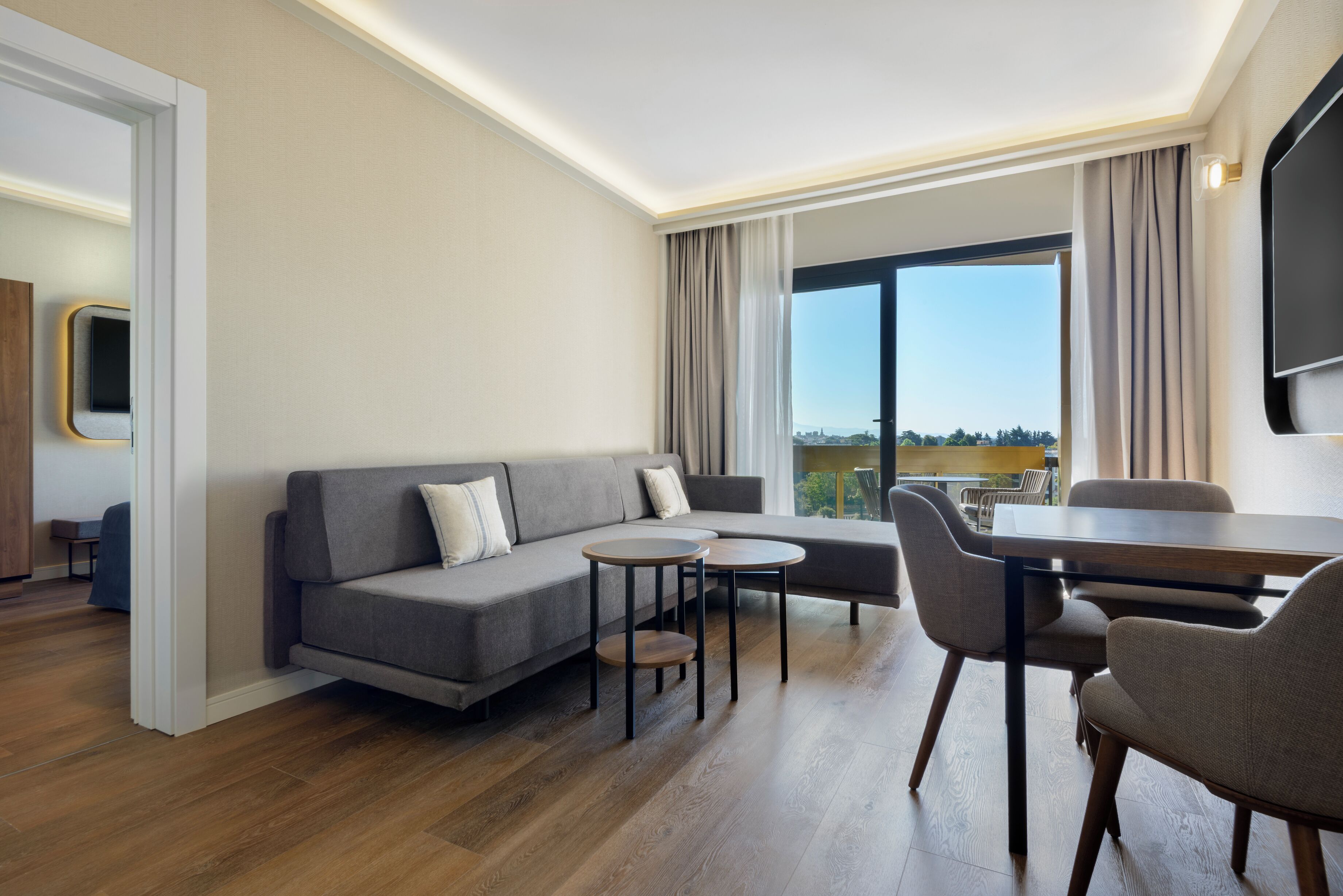 A modern hotel room with a grey sofa, a round coffee table, and a dining area with chairs and a table.