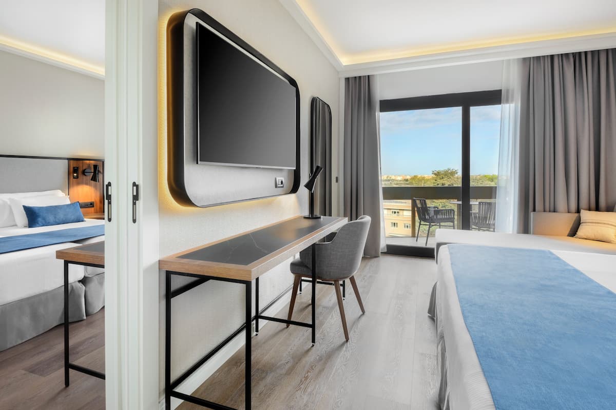 A modern hotel room with a large bed, a desk, a chair, a flat-screen TV, and a balcony with a view.