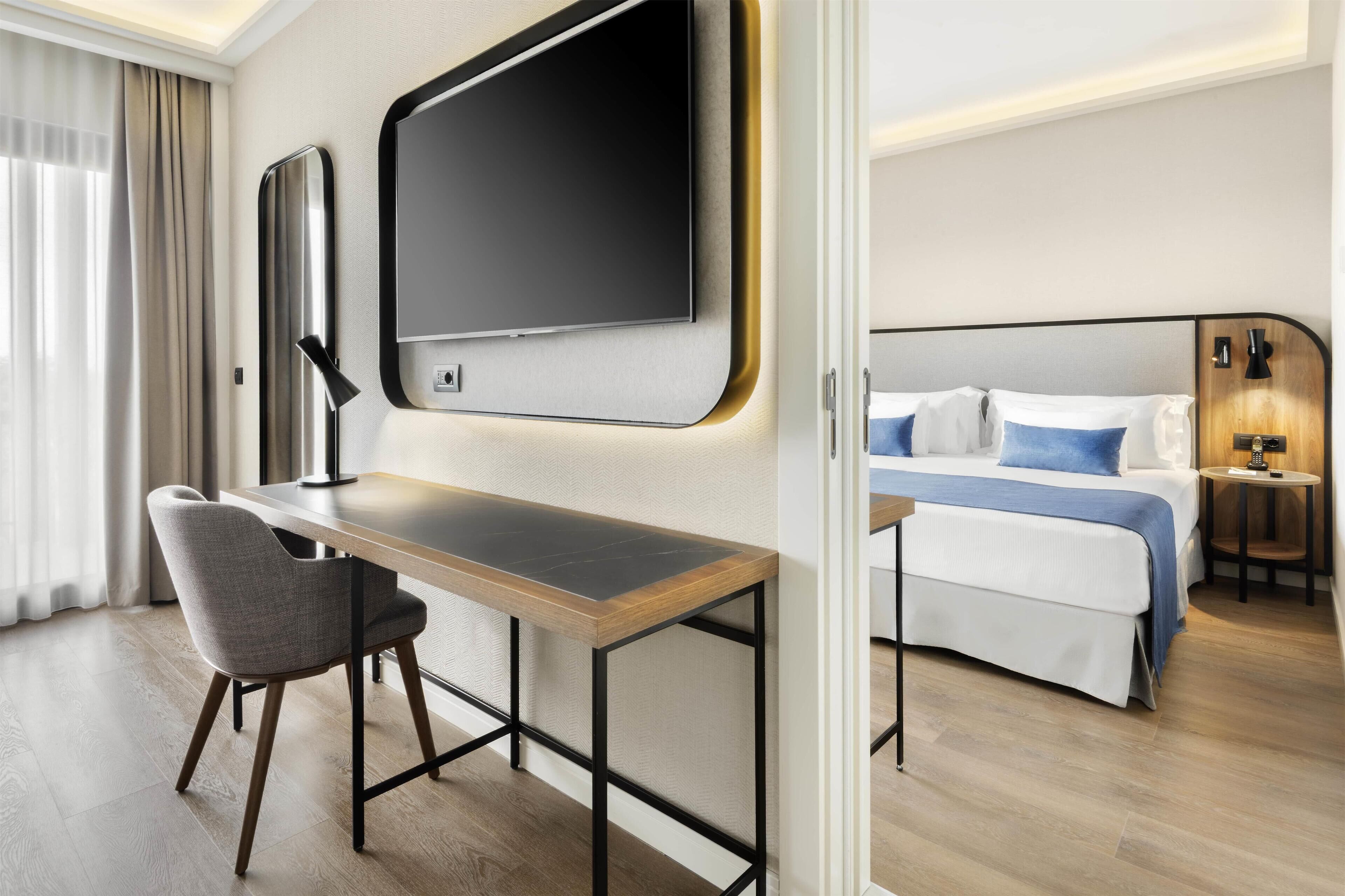 A hotel room with a bed, a desk, a chair, a TV, and a bedside table with a lamp.