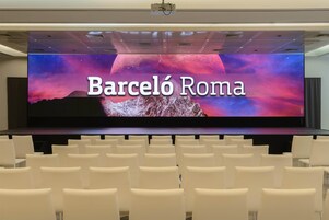 A conference room with a large screen displaying 'Barcelona Roma' and rows of white chairs.