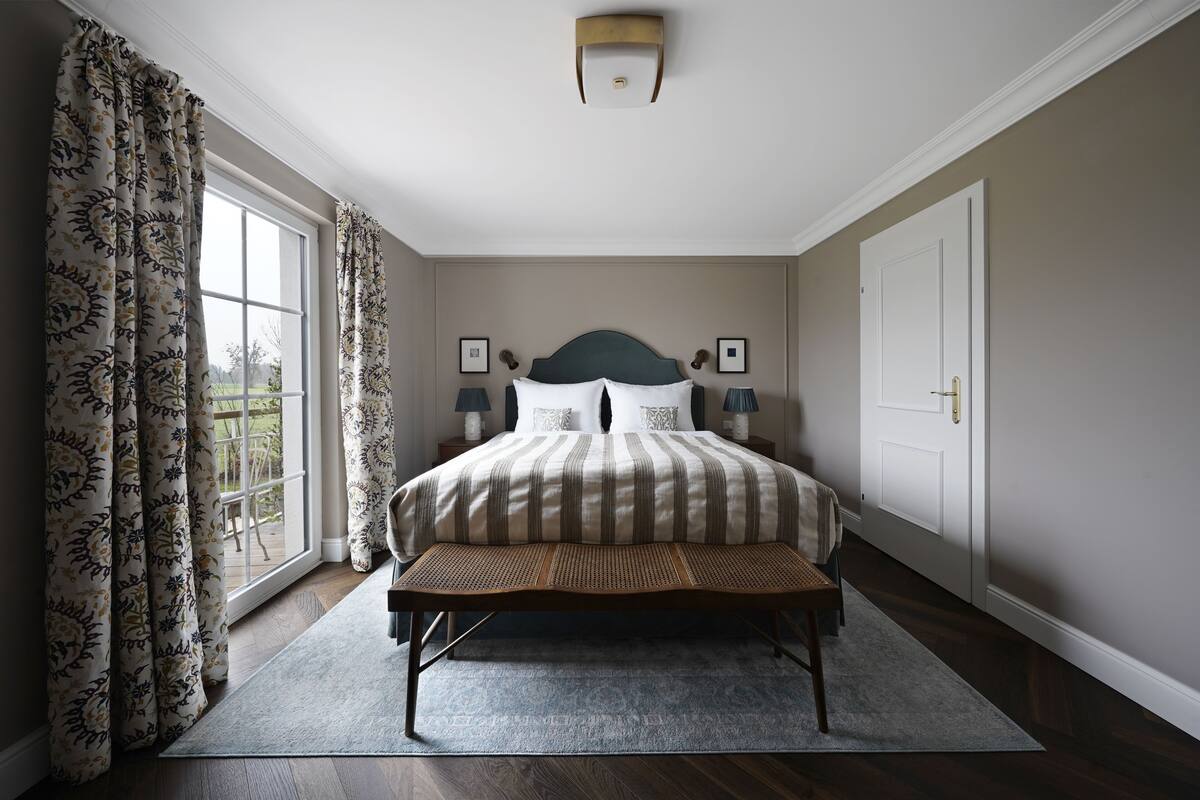 Um quarto com uma cama grande, um banco, uma janela com cortinas e uma porta.