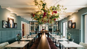 A restaurant with a long central bench, multiple tables, and chairs. There are framed artworks on the walls and a hanging floral arrangement above the central bench. - Numa Salzburg Ivy (Anif)