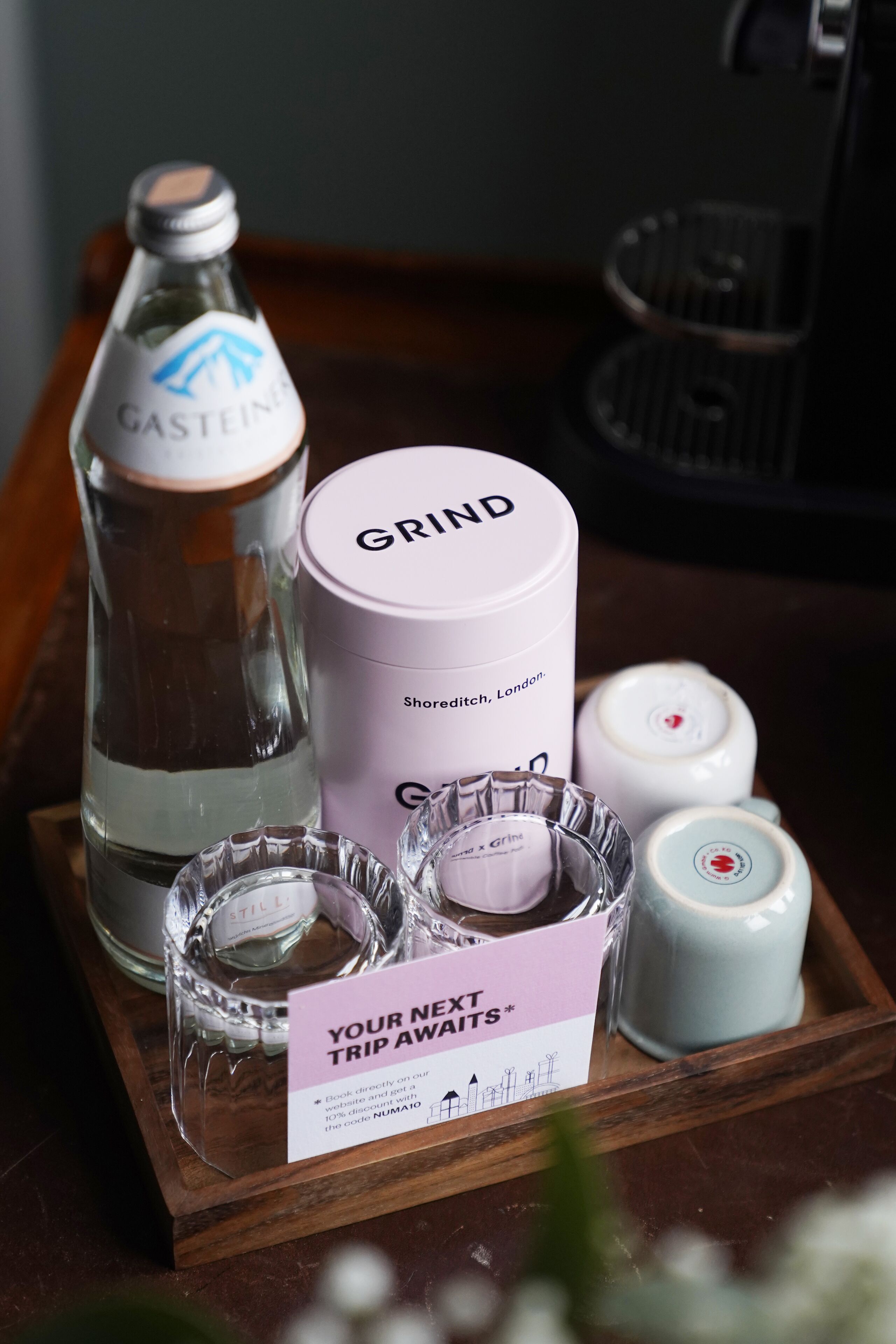 Un plateau contenant une bouteille de Gastein, un récipient blanc étiqueté « GRIND », deux petits récipients en verre et un panneau indiquant : « VOTRE PROCHAIN VOYAGE VOUS ATTEND ».