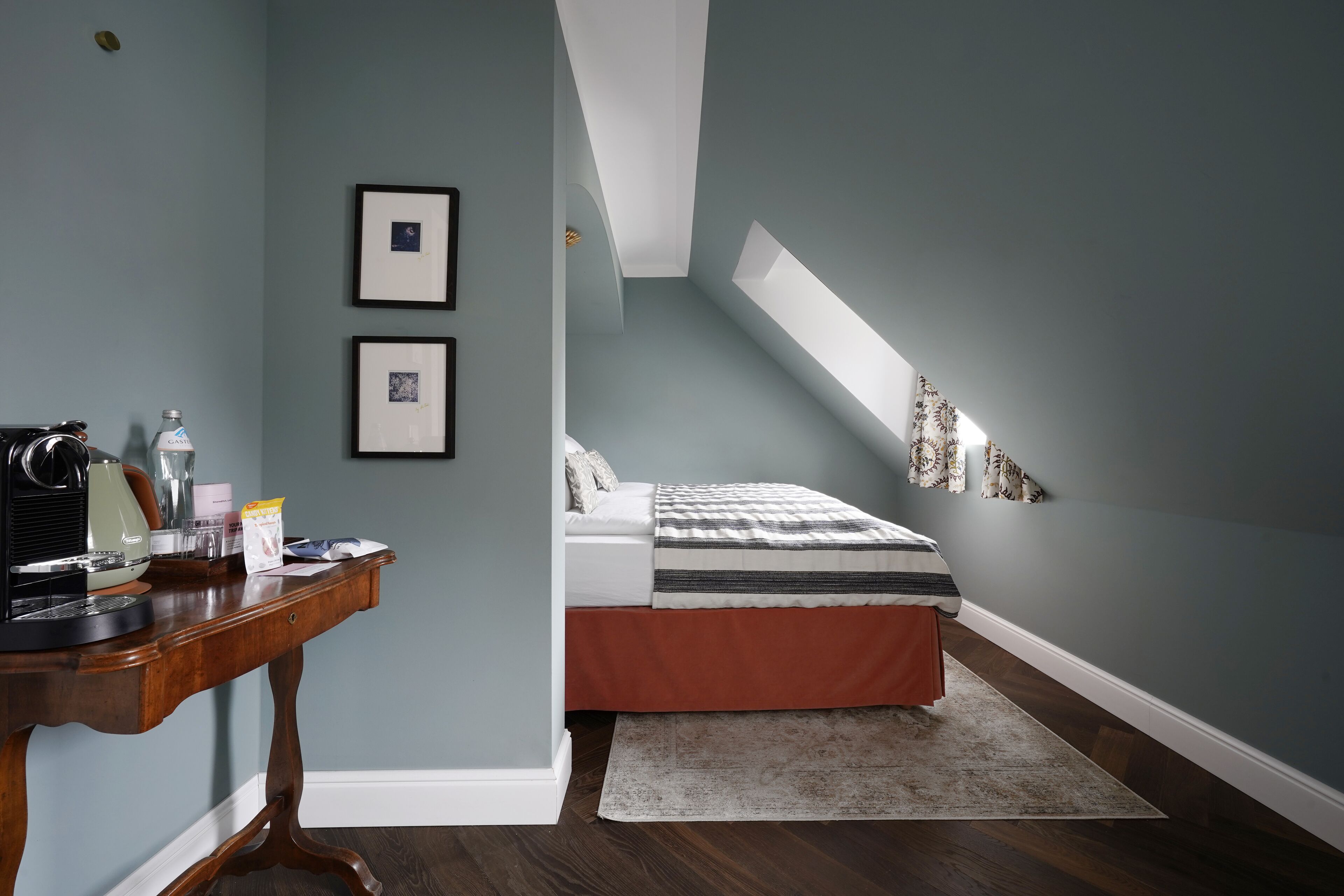 A bedroom with a sloped ceiling, a bed with a striped bedspread, a desk with a computer, and framed pictures on the wall.
