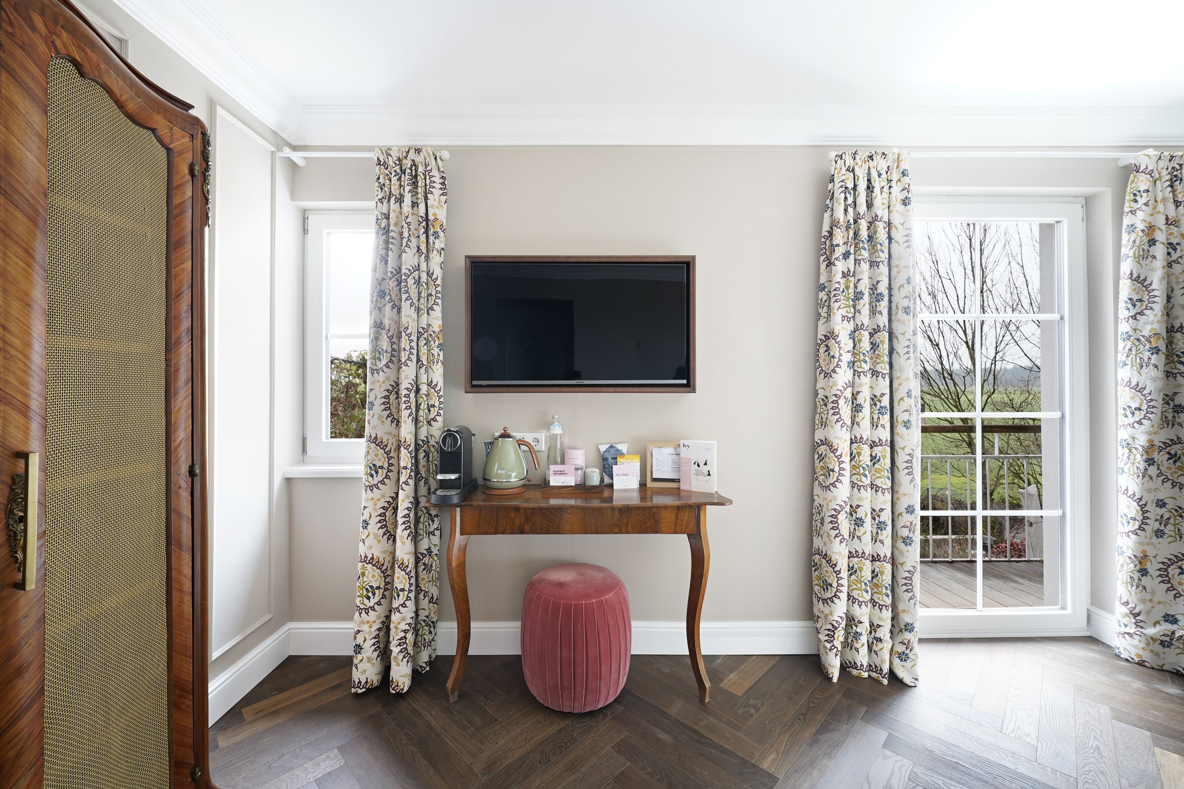 Ein Zimmer mit Schreibtisch aus Holz, einem Flachbildfernseher, einem rosa Hocker, gemusterten Vorhängen und Blick ins Freie.