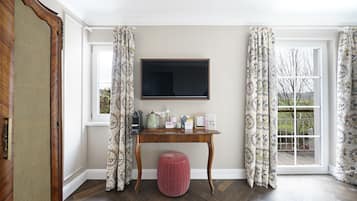 A room with a wooden desk, a flat-screen TV, a pink ottoman, patterned curtains, and a view of the outdoors.