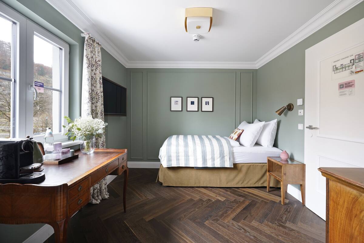 Quarto com cama, escrivaninha, cadeira, luminária e janela com cortinas.