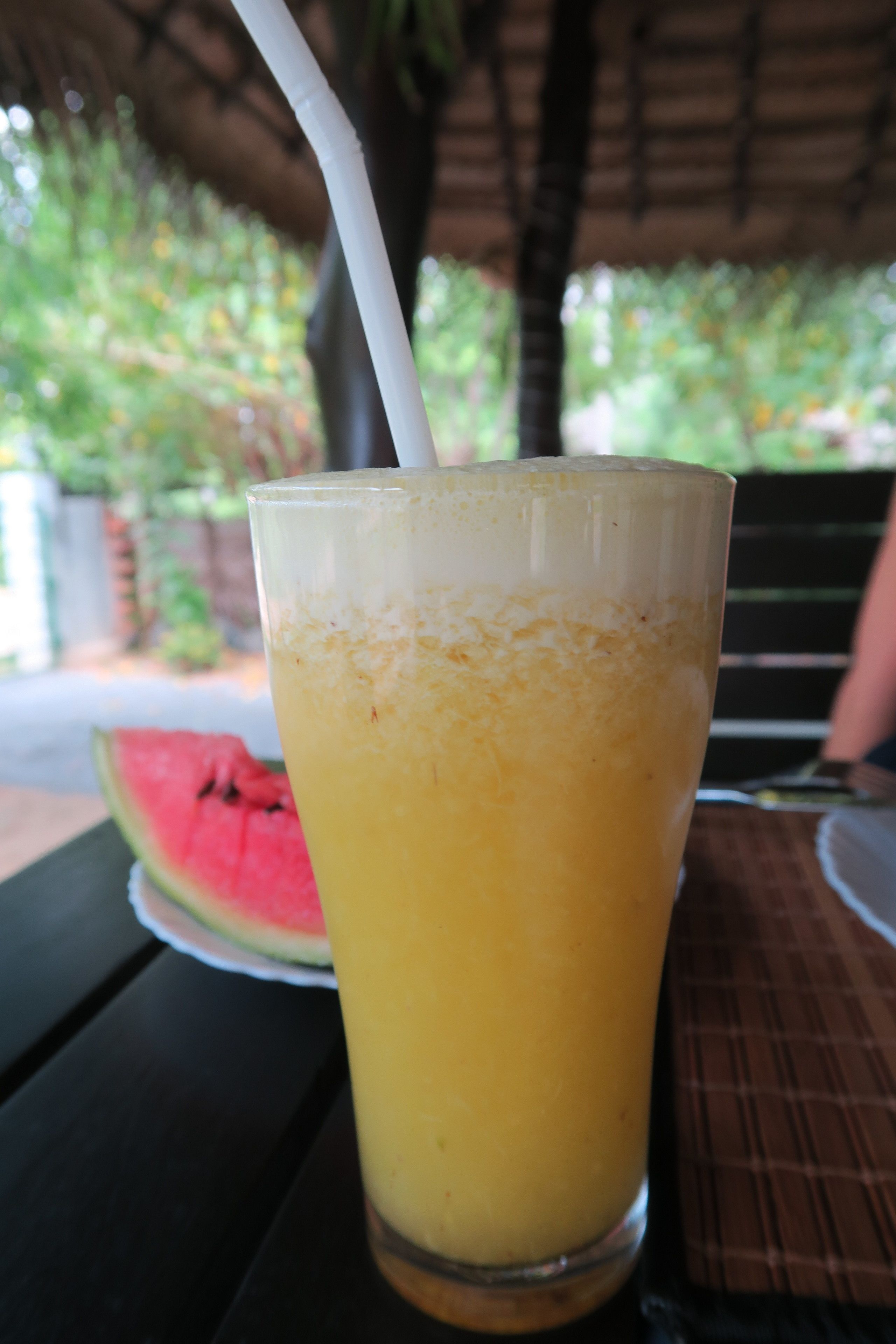 Un verre de jus d’orange avec une paille, posé sur une table, à côté d’une tranche de pastèque.