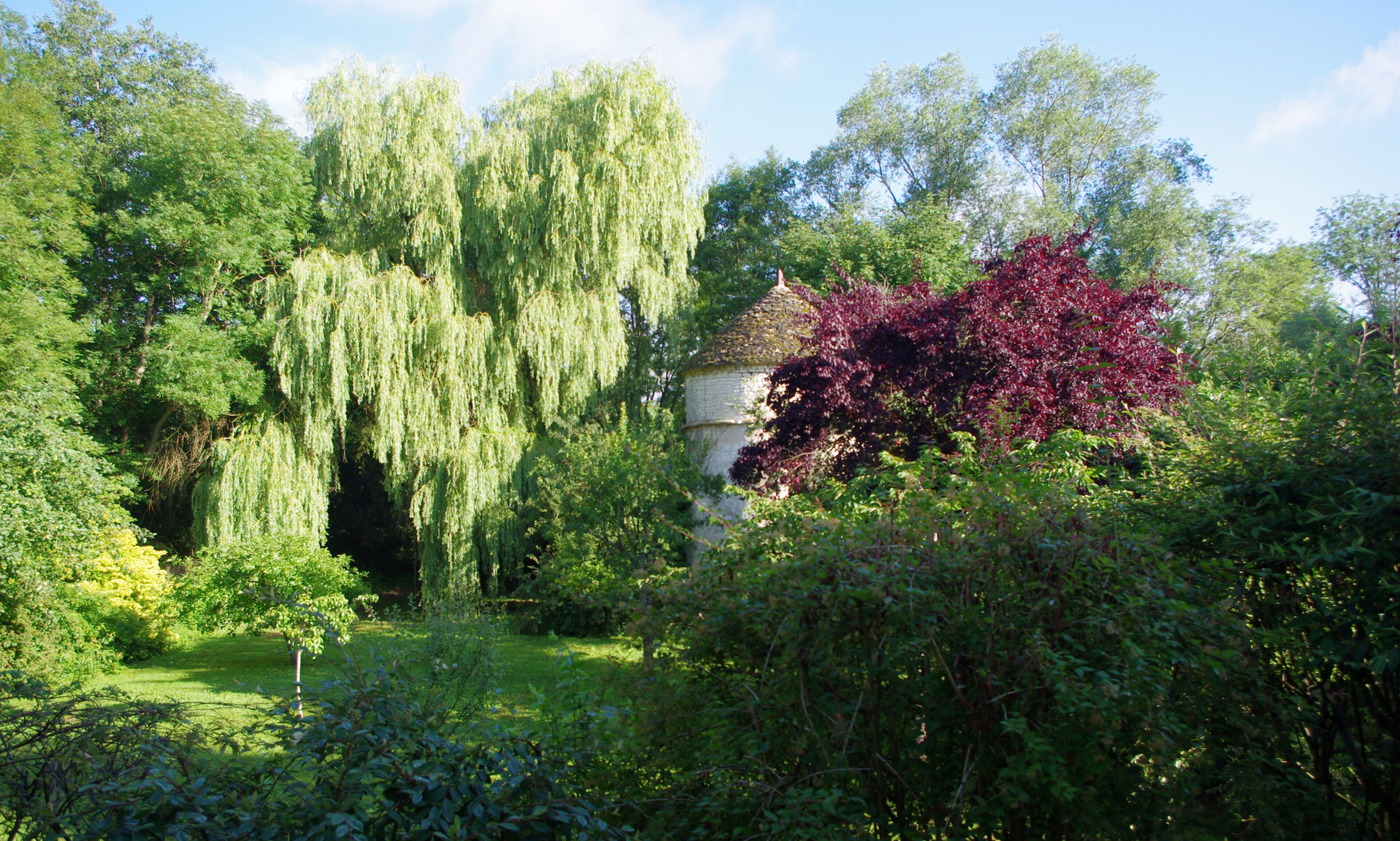 Een tuin met diverse bomen, waaronder huilwilgen en een boom met rode bladeren, en een klein wit gebouw met een puntig dak.
