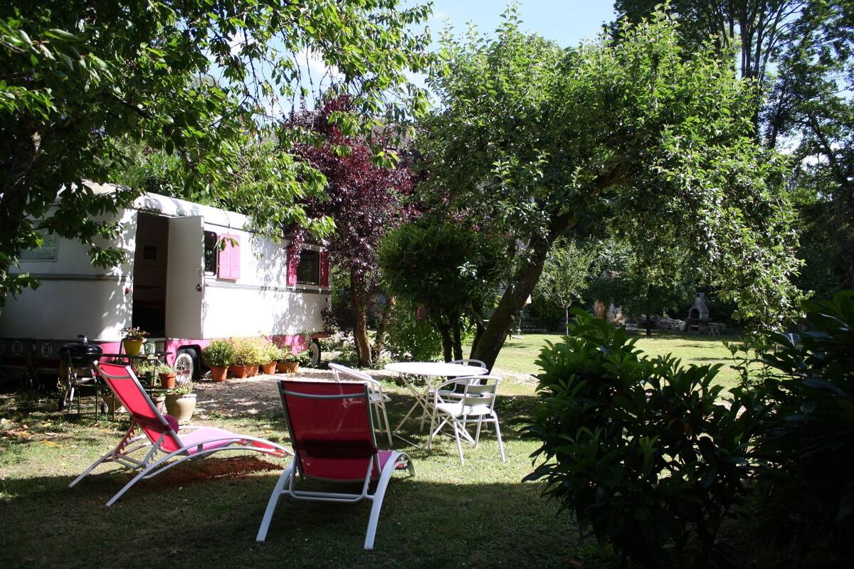 Un terrain de camping équipé d’une caravane, d’un espace extérieur aménagé pour s’asseoir et d’une végétation luxuriante.
