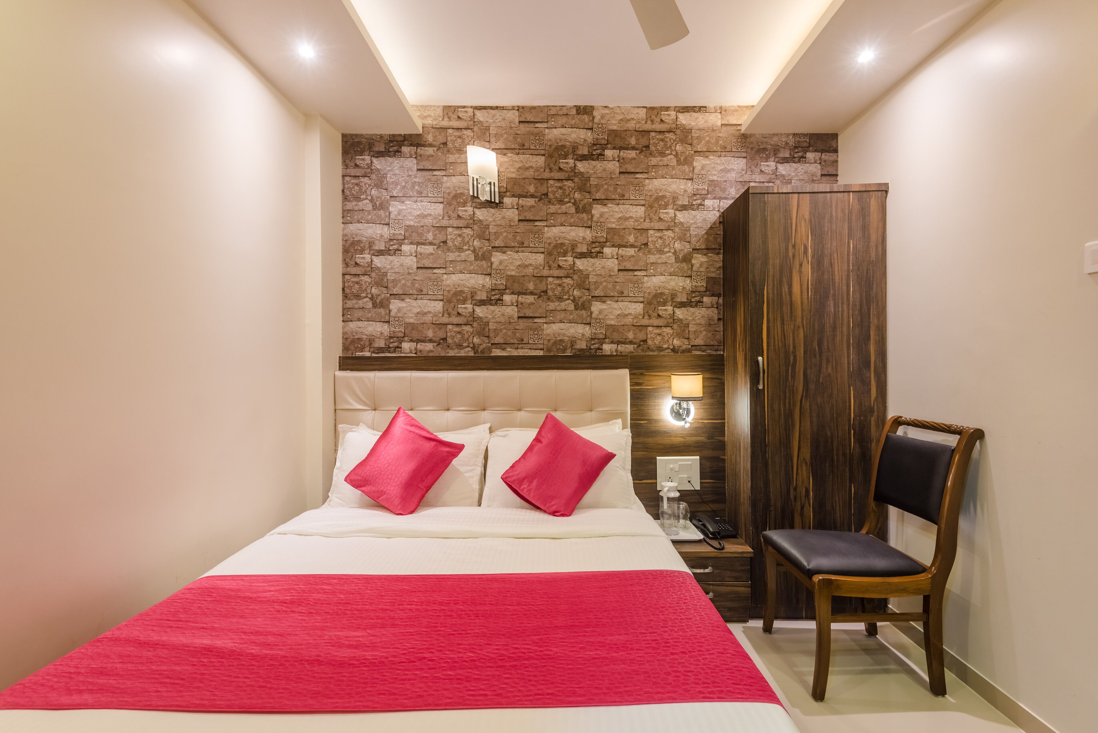 Een slaapkamer met een stenen muur, een bed met witte beddengoed en rode kussens, een houten garderobe en een stoel.