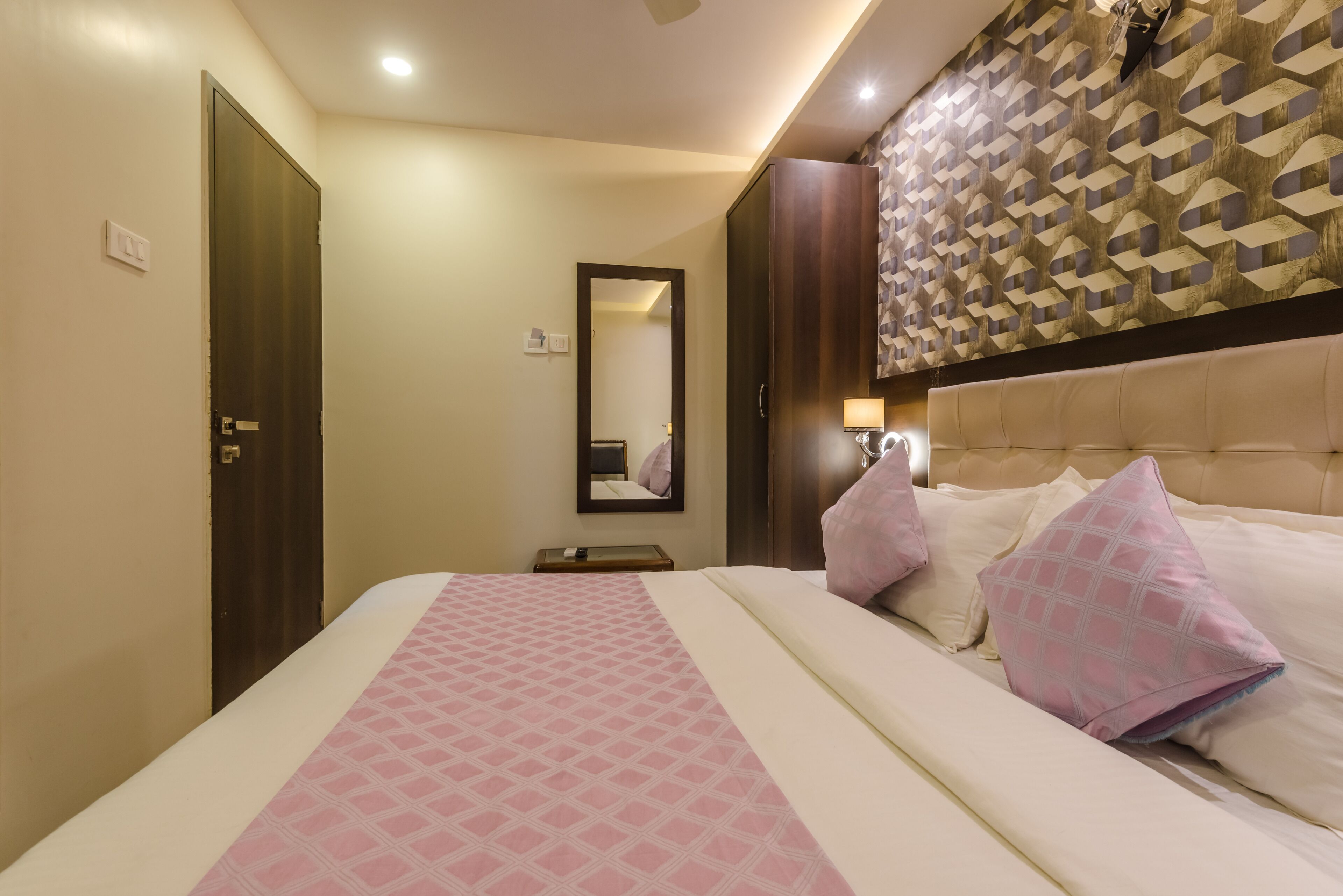 Habitación de hotel con cama, mesita de noche, espejo y un papel tapiz estampado.
