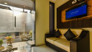 A modern living room with a beige sofa, a mounted TV displaying 'Vivo Smart Phone', and a glass door leading to an outdoor area with potted plants.
