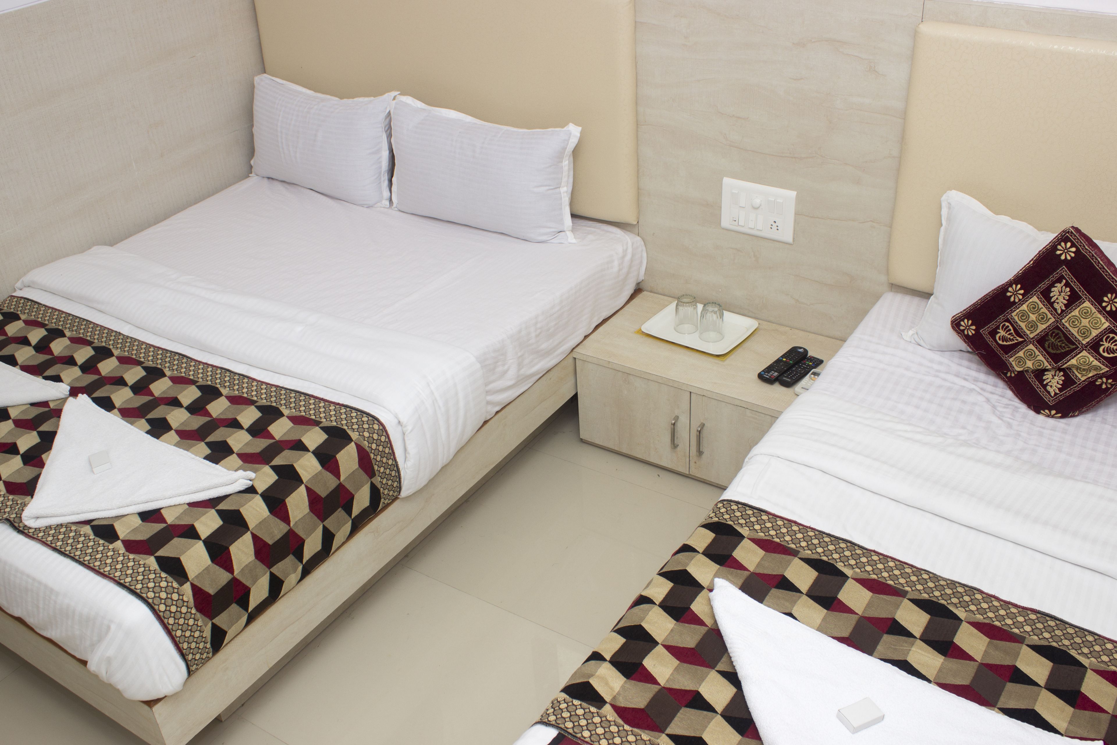 Two single beds with white linens, a patterned bedspread, a bedside table with a remote control, and a decorative pillow.