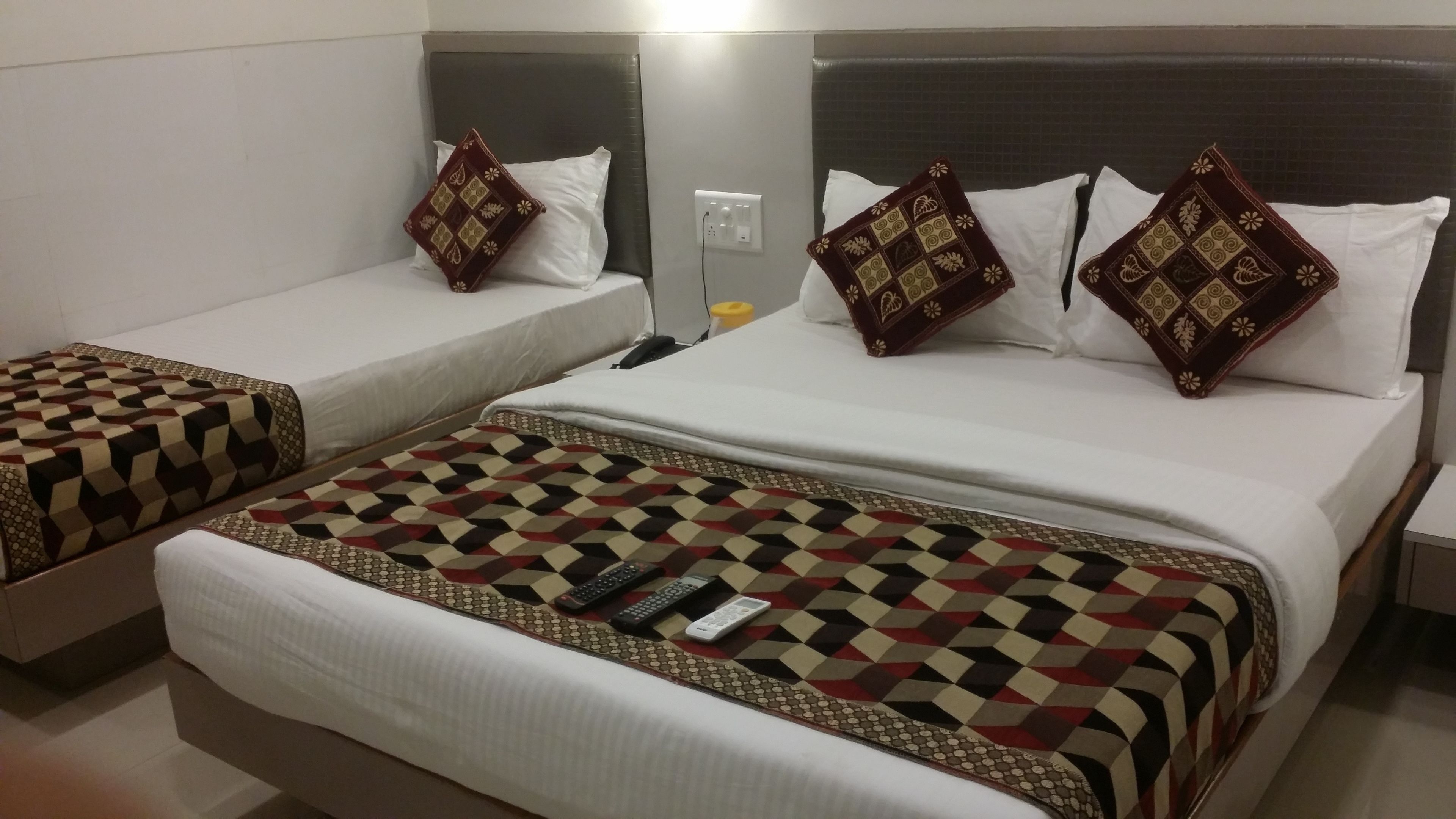 A hotel room with two single beds, each with a patterned bedspread and matching pillows. There is a larger bed with a similar patterned bedspread and pillows. A remote control and a phone are placed on the larger bed.