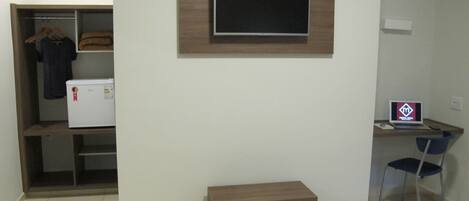 A bedroom with a bed, a desk with a laptop, a chair, a small wooden bench, a wardrobe, and a refrigerator.