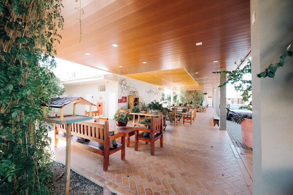 Un espacio interior amplio con mesas y sillas de madera, plantas en macetas y piso de baldosas.