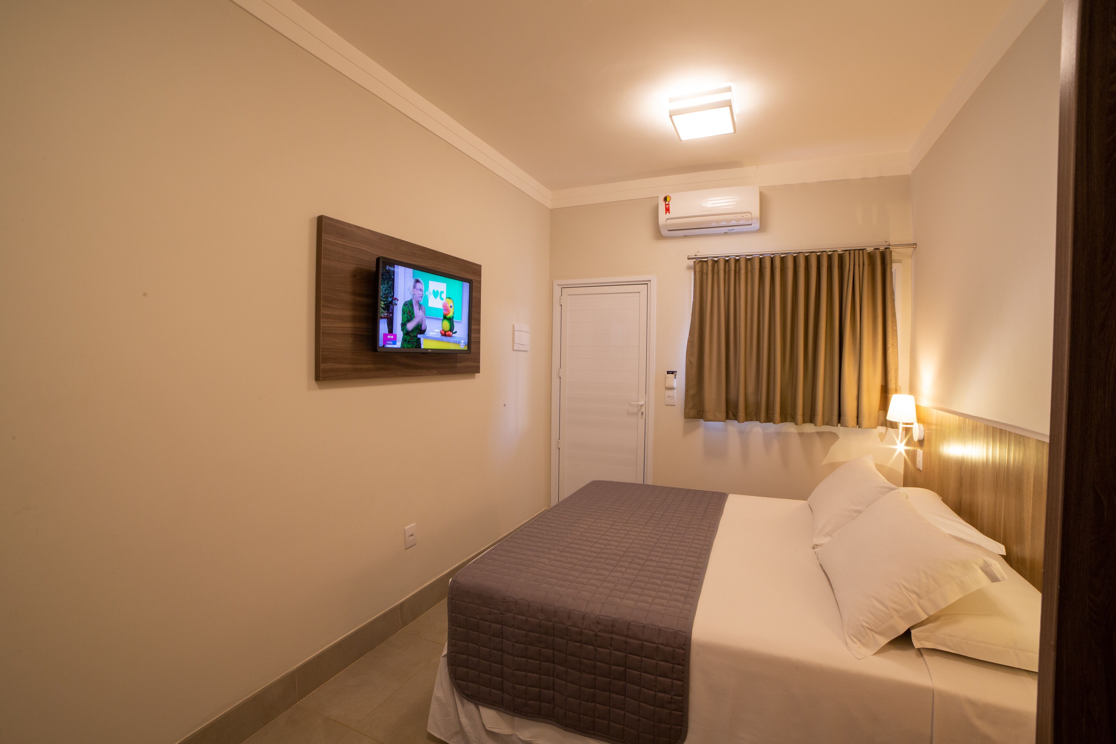 Habitación de hotel con cama, televisor montado en la pared, una puerta y una ventana con cortinas.