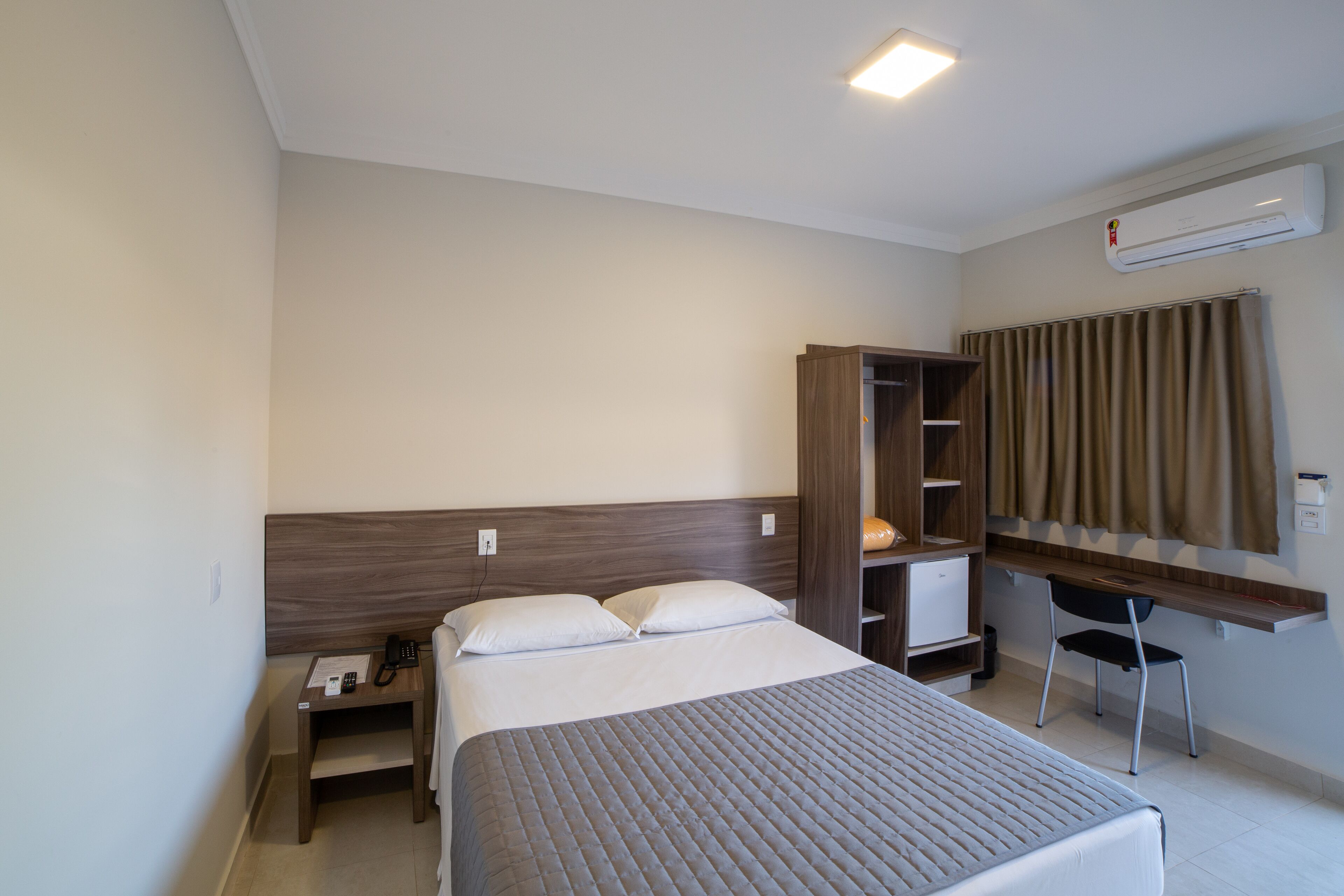 Habitación de hotel con cama, mesita de noche, escritorio, silla, armario y equipo de aire acondicionado.