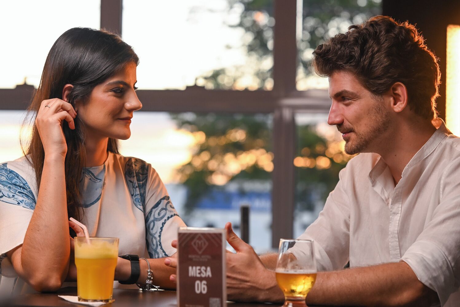 Deux personnes sont assises à une table, devant des boissons. On peut voir un panneau indiquant « MESA 06 » et une fenêtre donnant sur des arbres.