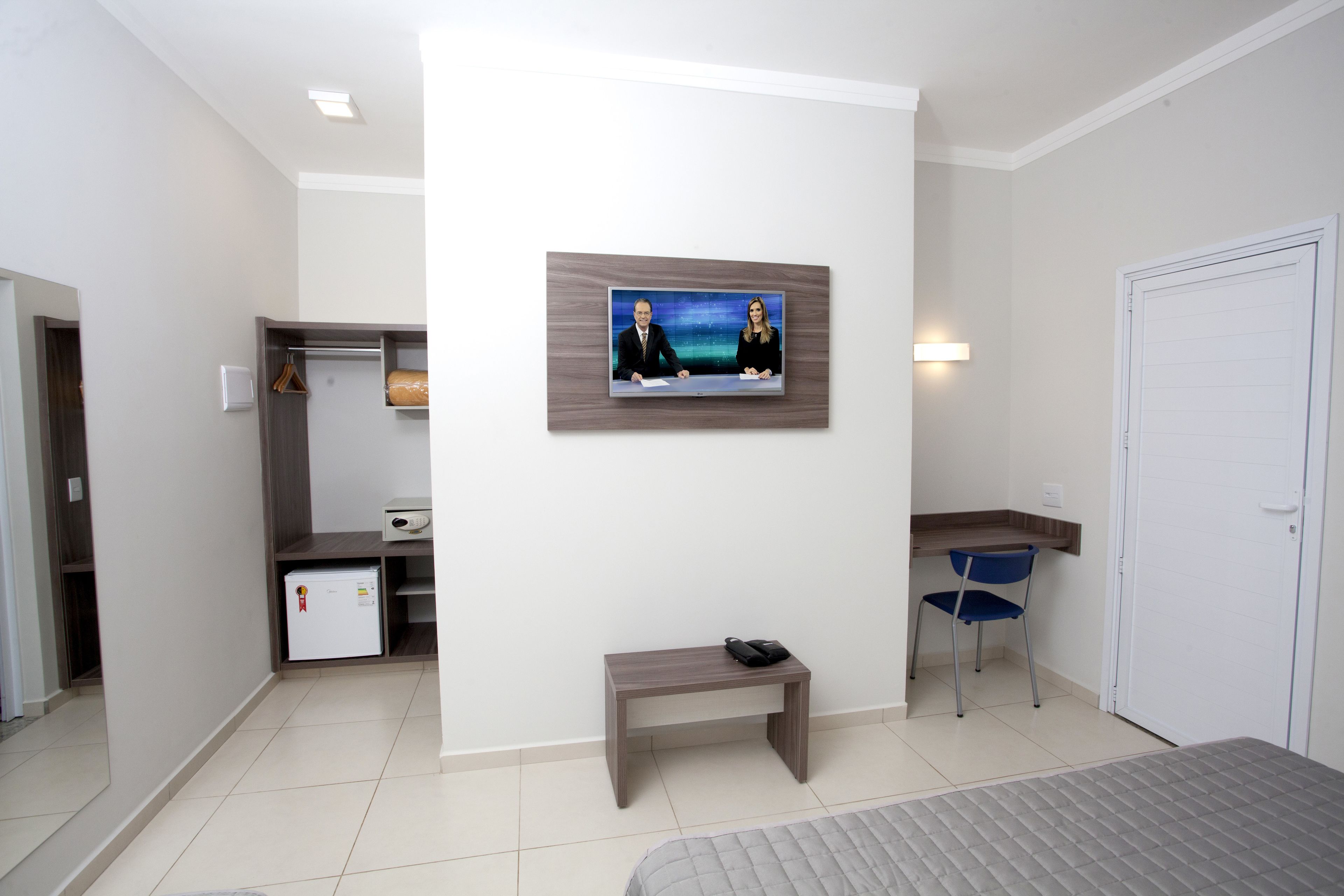 Una habitación de hotel con cama, escritorio, silla, mesita, televisor montado en la pared y armario.