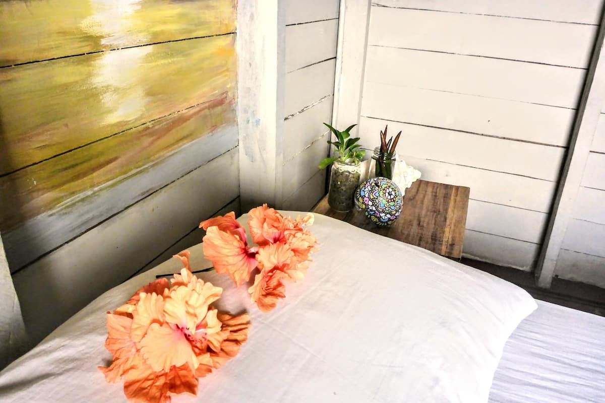 Ein Bett mit weißer Bettdecke, einem Holzkopfstück und einer Blumenkranz. Auf einem Holznachttisch stehen zwei kleine Blumentöpfe.