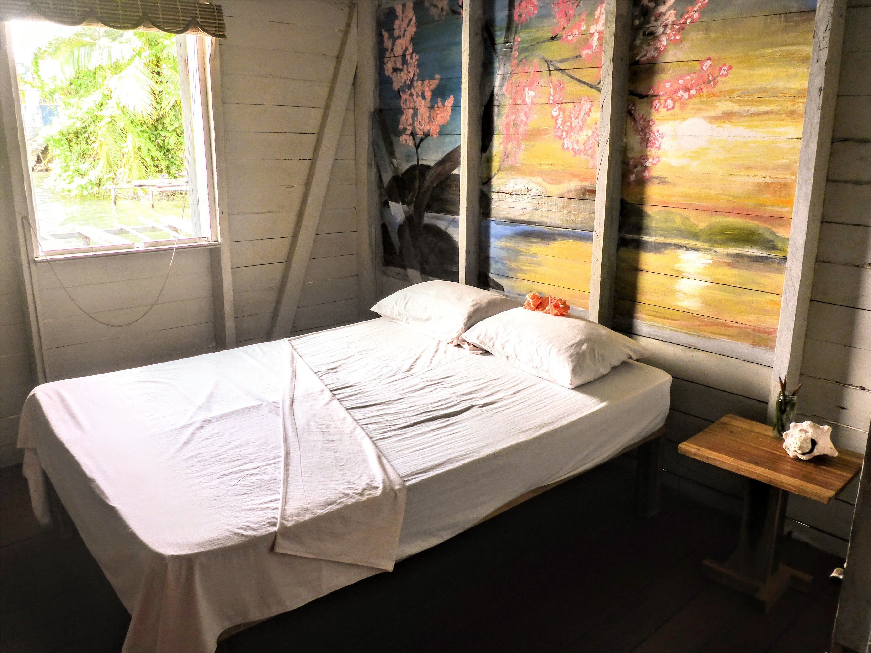 A neatly made bed with white linens, a small bedside table with a vase, and a window with a view of greenery.